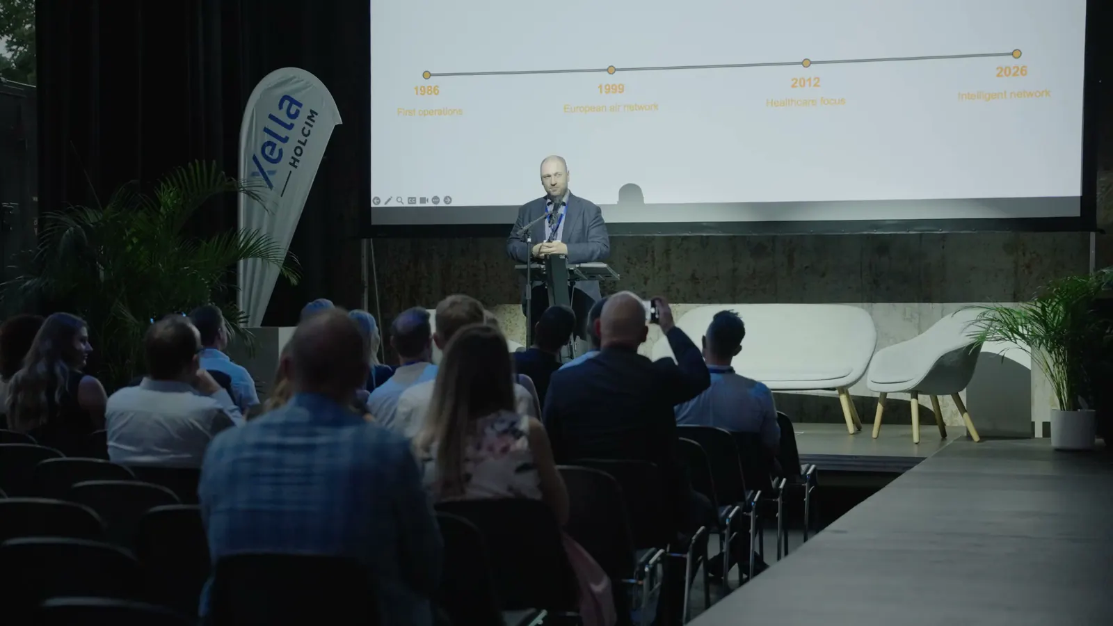 Referent am Rednerpult vor einer Zeitleiste auf der Projektionswand, davor das sitzende Publikum in der Voltahalle