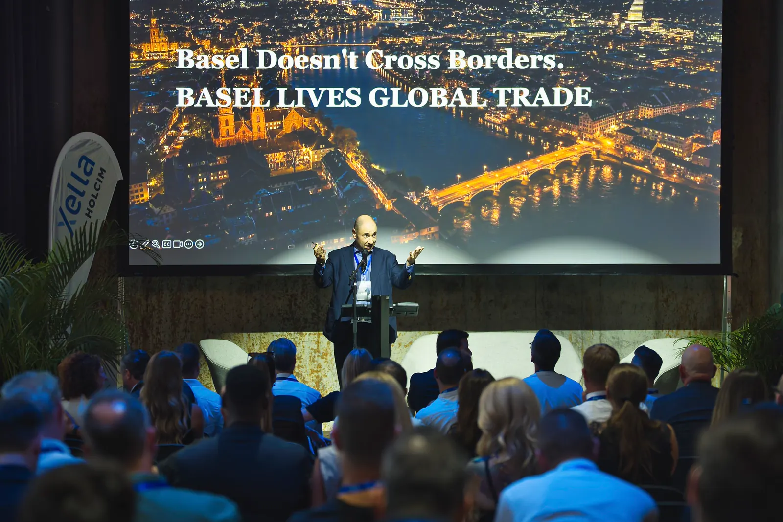 Referat auf der Bühne vor vollem Saal, auf der Leinwand steht Basel Lives Global Trade